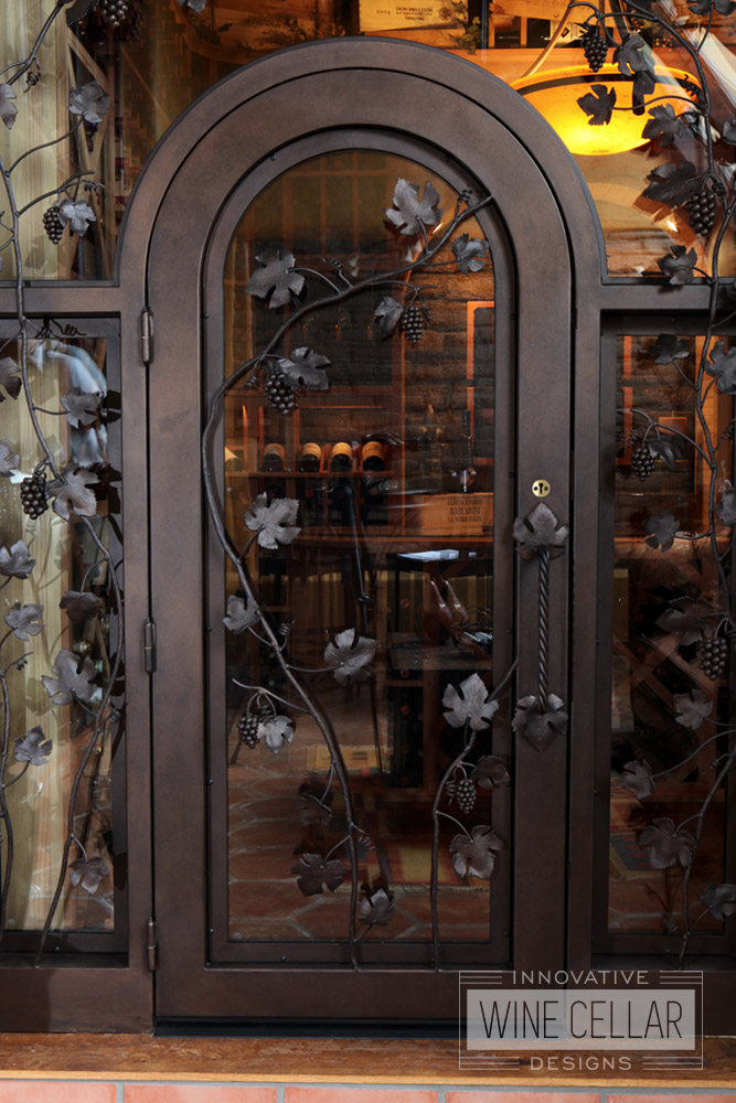 Custom Glass Wine Cellar Door Design with Wrought Iron Archway and Grape Vine Accents by Innovative Wine Cellar Designs.
